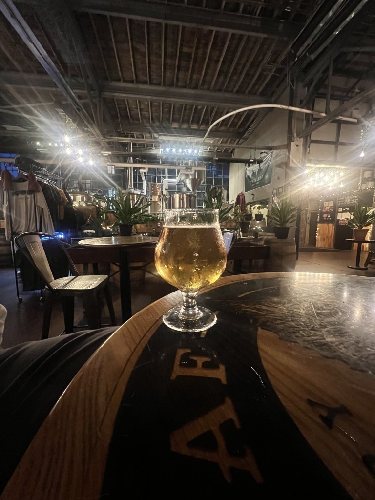 A glass of golden beer on a wooden tabletop in a brewery or bar setting, with dim lighting and industrial decor in the background.