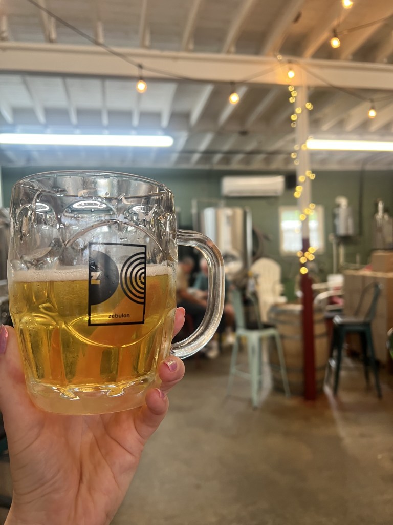 A hand holding a clear beer mug filled with golden beer, inside a brewery with a blurred background featuring brewing equipment and warm string lights.