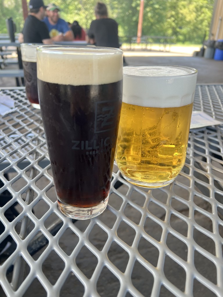 Two glasses of beer on a metal table; one is dark with a creamy head and the other is light and bubbly.