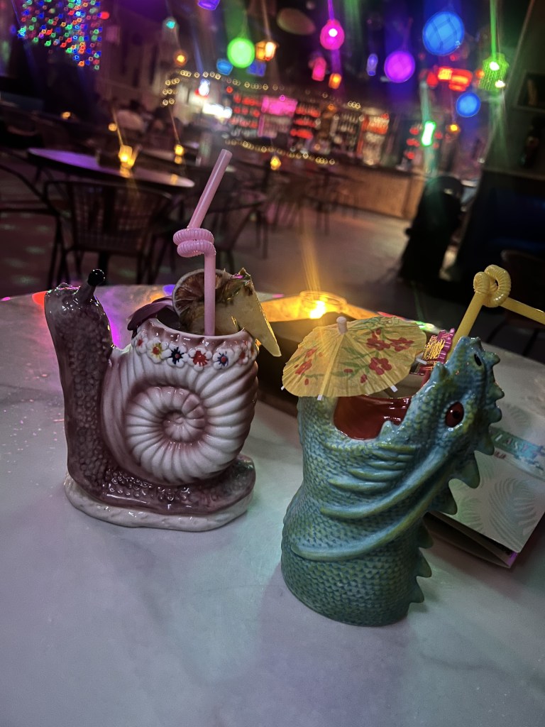 Two whimsical cocktail mugs shaped like a snail and a dragon, each decorated with colorful garnishes and straws, set on a table in a dimly lit bar with vibrant lights in the background.