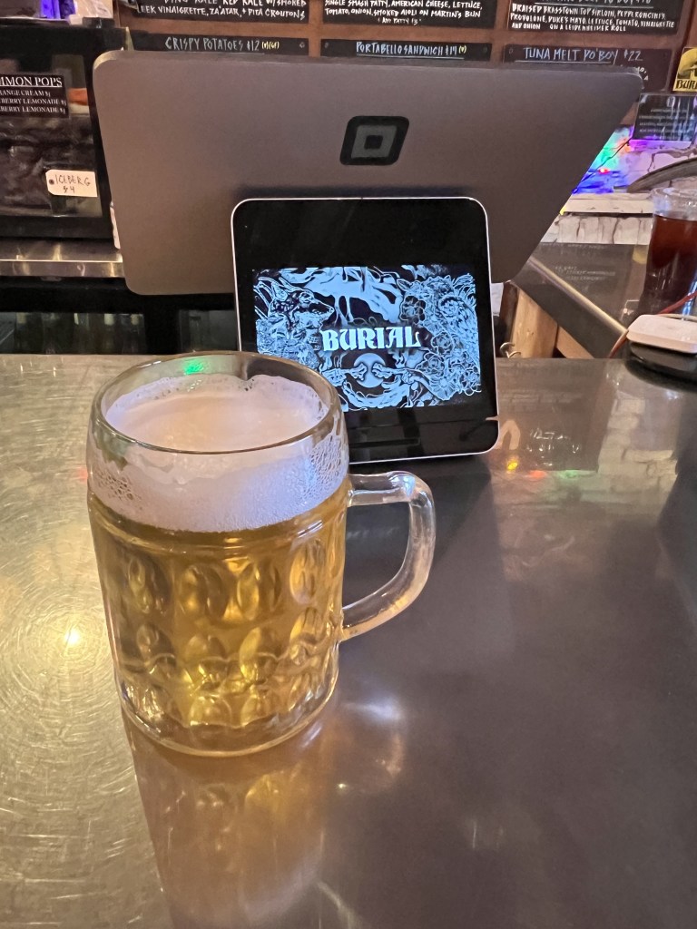 A frosty beer mug filled with light beer sits on a bar counter, with a tablet displaying the word 'BURIAL' in stylized lettering in the background.