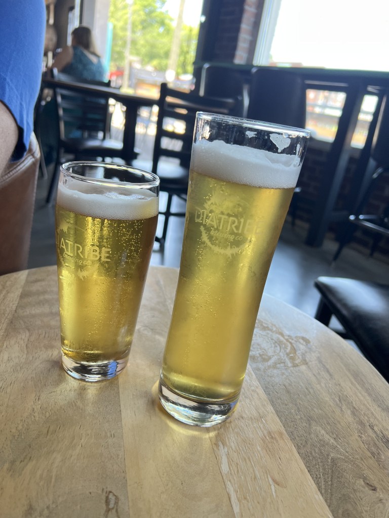Two glasses of beer on a wooden table in a bar setting, one tall and one short, with a foamy head.
