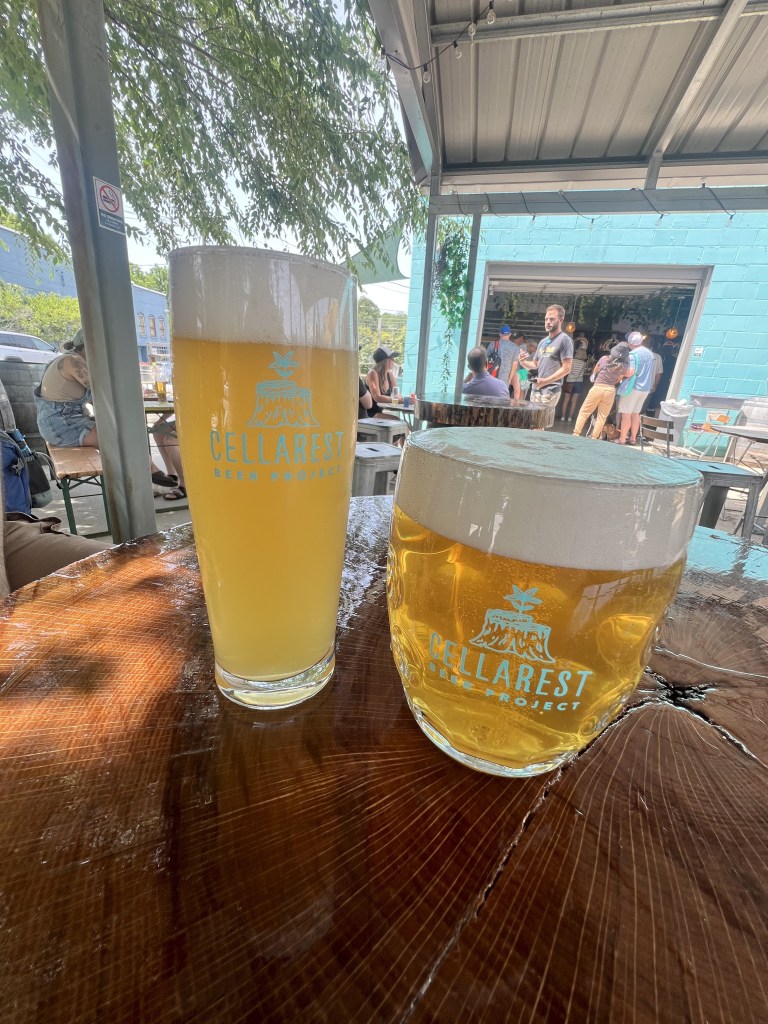 Two glasses of beer on a wooden table at an outdoor setting, featuring the logo of 'Cellarest Beer Project'.