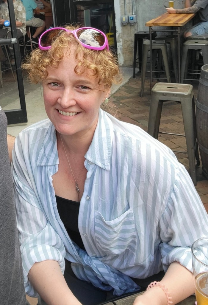 A smiling woman with curly hair, wearing a blue and white striped shirt and pink sunglasses, sitting at a table in an outdoor setting.